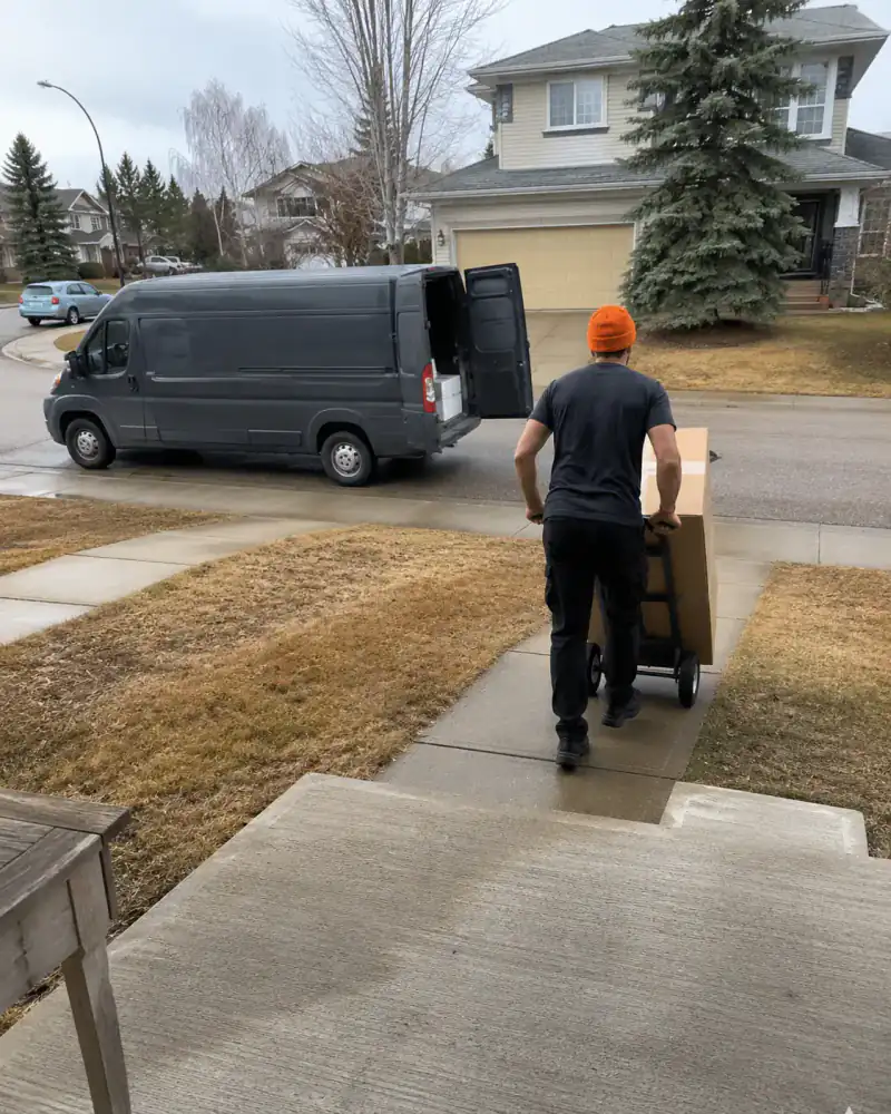 Mover wheeling a large boxed delivery up a front walk from the open cargo van at the curb